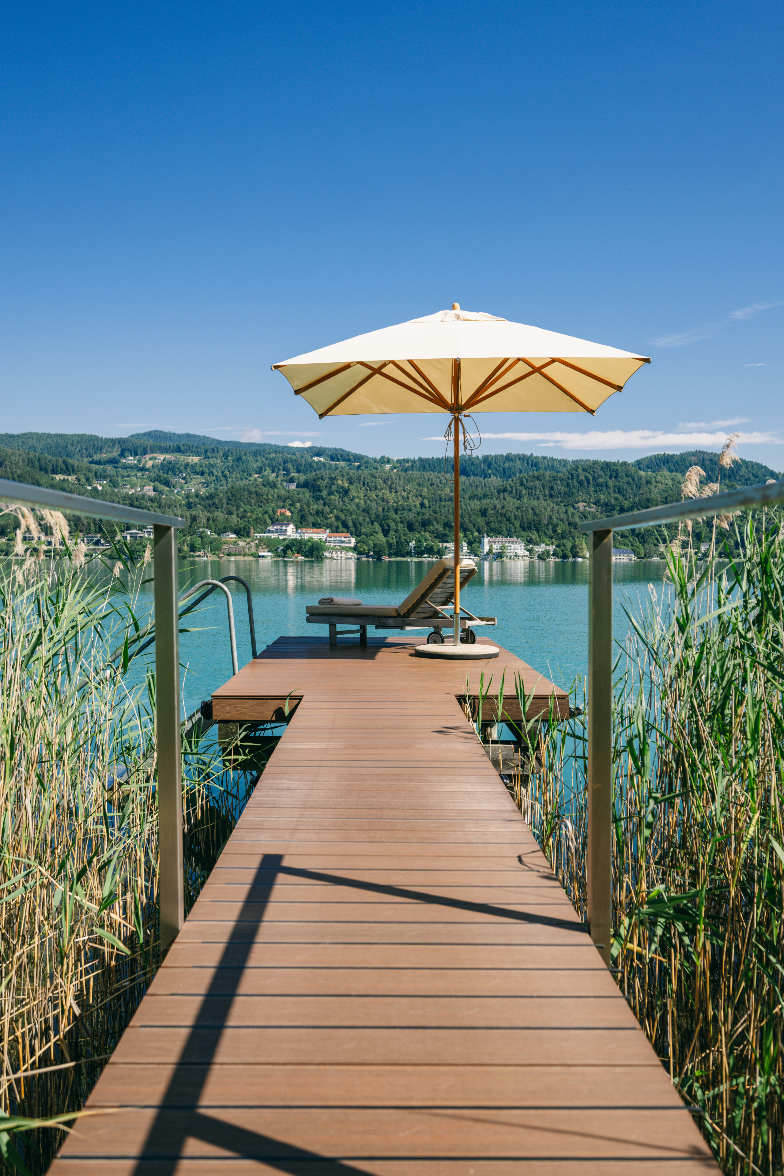 Ein sonniger Ausblick von einer Holzterrasse auf einem ruhigen See, mit einem Liegestuhl und einem Sonnenschirm.